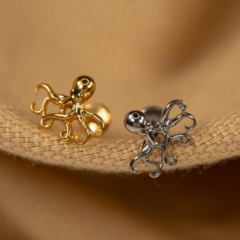 May include: Two octopus earrings, one gold and one silver, with a detailed design. The earrings are on a brown fabric background.