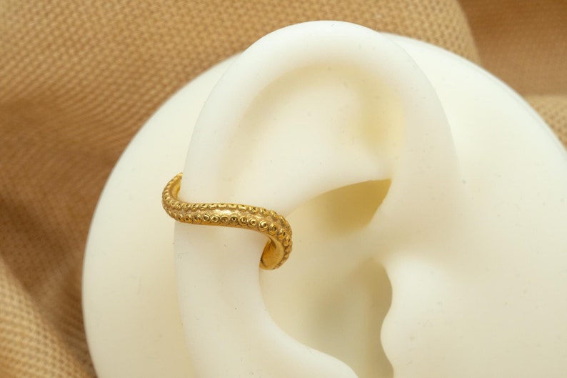 May include: A gold ear cuff with a textured, beaded design. The cuff is curved and has a simple, minimalist style.