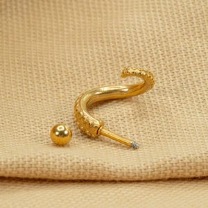 May include: Gold-toned body jewelry featuring a curved, textured bar with a small ball at the end. The bar is designed to resemble an octopus tentacle.