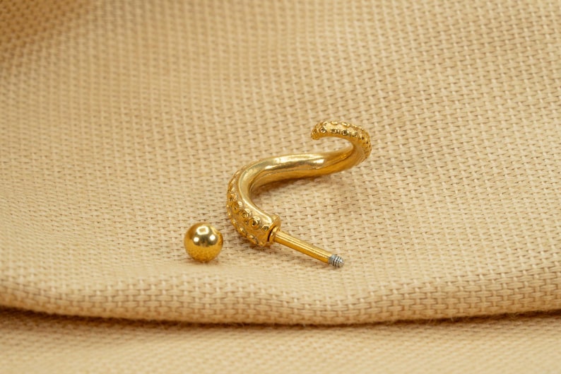 May include: Gold-toned body jewelry featuring a curved, textured bar with a small ball at the end. The bar is designed to resemble an octopus tentacle.