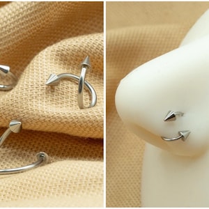 May include: Two silver nose rings with pointed ends. The rings are twisted and have a curved shape. One ring is shown on a model's nose.