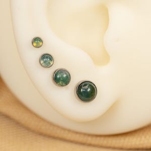 Puede incluir: Cuatro pendientes de &oacute;palo verde y blanco de diferentes tama&ntilde;os, engastados en plata, usados en la oreja de una persona.
