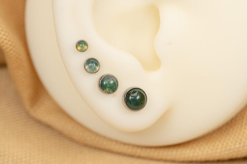 Puede incluir: Cuatro pendientes de &oacute;palo verde y blanco de diferentes tama&ntilde;os, engastados en plata, usados en la oreja de una persona.