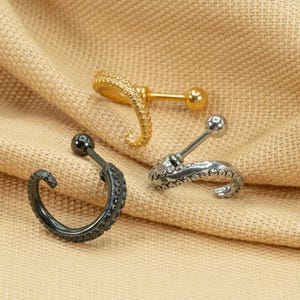 May include: Three different styles of octopus-shaped cartilage earrings. The earrings are made of silver, gold, and black metal. The earrings are on a beige fabric background.