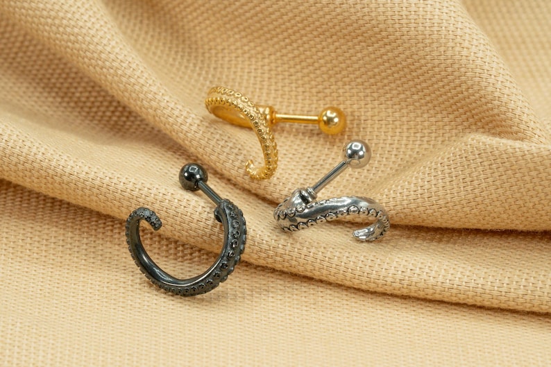 May include: Three different styles of octopus-shaped cartilage earrings. The earrings are made of silver, gold, and black metal. The earrings are on a beige fabric background.