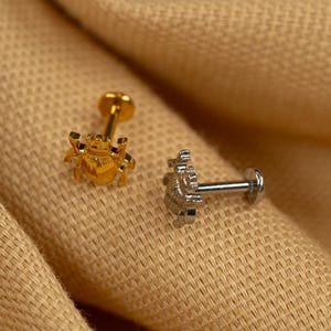 Puede incluir: Dos peque&ntilde;os piercings corporales decorativos. Uno es dorado y presenta un dise&ntilde;o de escarabajo. El otro es plateado y presenta un dise&ntilde;o floral.