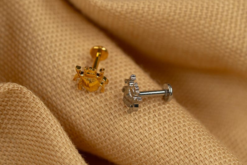 Puede incluir: Dos peque&ntilde;os piercings corporales decorativos. Uno es dorado y presenta un dise&ntilde;o de escarabajo. El otro es plateado y presenta un dise&ntilde;o floral.