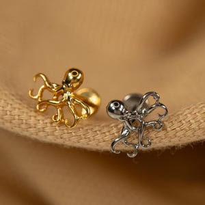 May include: Two octopus shaped earrings, one gold and one silver, on a brown fabric background.