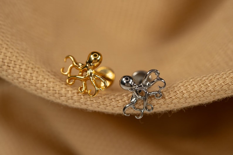 May include: Two octopus shaped earrings, one gold and one silver, on a brown fabric background.