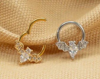 Puede incluir: Dos anillos de septum plateados y dorados con un diseño de murciélago y brillantes transparentes. Los anillos están sobre un fondo de tela beige.