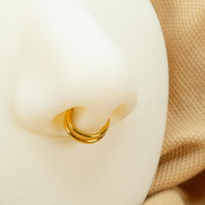以下が含まれることがあります： 2 つの金のフープで鼻のピアスをクローズアップ。フープは互いに重ねられており、シンプルでクラシックなデザインです。