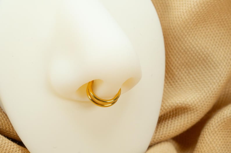 以下が含まれることがあります： 2 つの金のフープで鼻のピアスをクローズアップ。フープは互いに重ねられており、シンプルでクラシックなデザインです。