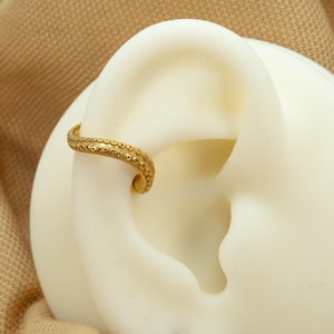 May include: Gold ear cuff with a textured, beaded design. The cuff is curved and has a small, round bead at the end.