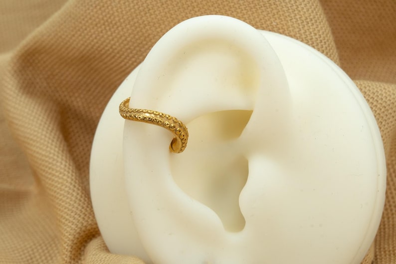 May include: Gold ear cuff with a textured, beaded design. The cuff is curved and has a small, round bead at the end.