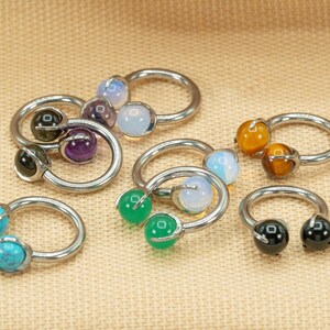 May include: A set of eight silver horseshoe rings with different colored gemstones. The gemstones include turquoise, green, black, brown, purple, and white.