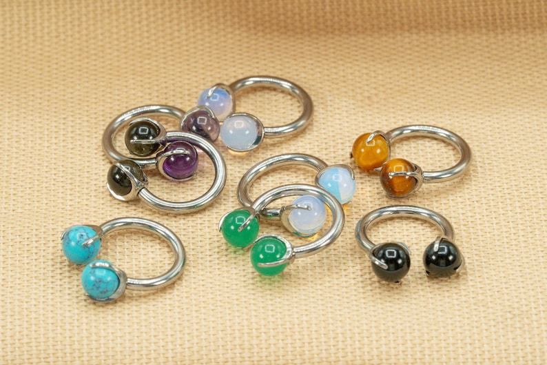 May include: A set of eight silver horseshoe rings with different colored gemstones. The gemstones include turquoise, green, black, brown, purple, and white.