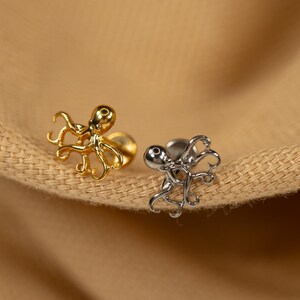 May include: Two octopus shaped earrings, one gold and one silver, on a brown fabric background.