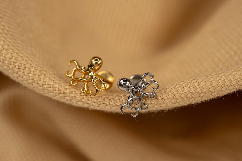 May include: Two octopus shaped earrings, one gold and one silver, on a brown fabric background.