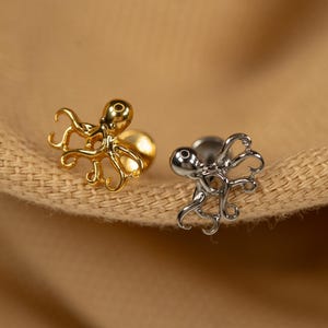 May include: Two octopus shaped earrings, one gold and one silver, on a brown fabric background.