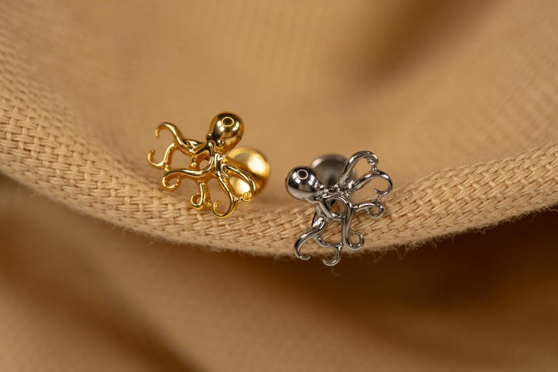 May include: Two octopus shaped earrings, one gold and one silver, on a brown fabric background.
