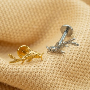 Op de afbeelding: Twee lichaamssieraden van goud en zilver met een vis- en hertontwerp. De sieraden liggen op een beige stoffen achtergrond.