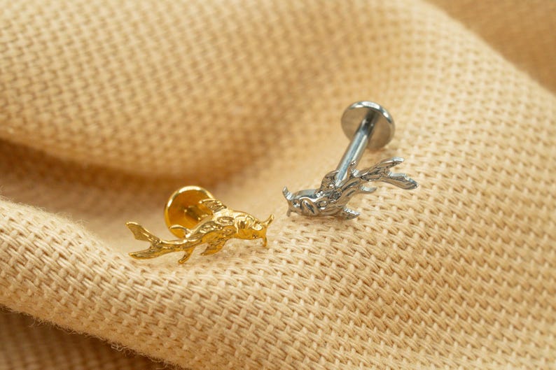 Op de afbeelding: Twee lichaamssieraden van goud en zilver met een vis- en hertontwerp. De sieraden liggen op een beige stoffen achtergrond.