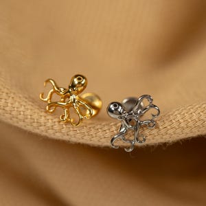 May include: Two octopus shaped earrings, one gold and one silver, on a brown fabric background.