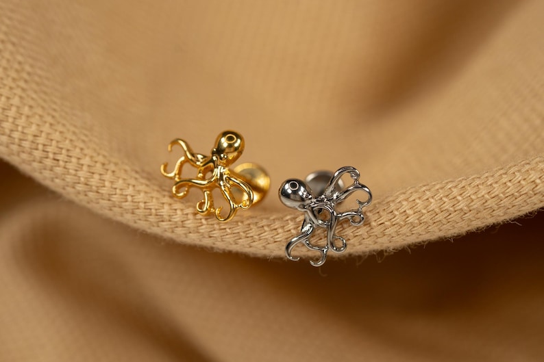 May include: Two octopus shaped earrings, one gold and one silver, on a brown fabric background.