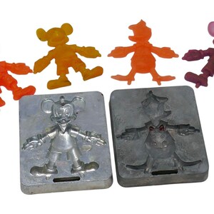May include: A collection of colorful, translucent cartoon character figures in orange, yellow, and maroon, along with two metal molds. The molds feature detailed character designs, likely for creating the figures. The figures are in various poses.