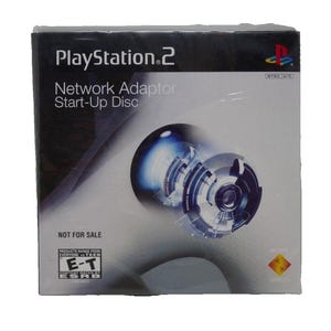 Puede incluir: Una caja de disco de inicio del adaptador de red PlayStation 2. La caja es en blanco y negro con el logotipo de PlayStation 2 y el texto "Network Adaptor Start-Up Disc". La caja también tiene la clasificación ESRB "E-T".