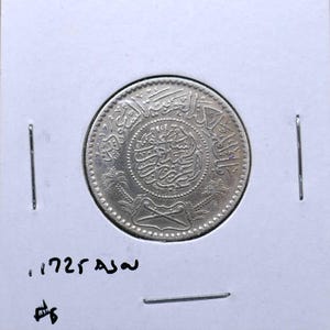 May include: A silver coin with intricate Arabic script and a central design. The coin is set in a white holder with vertical slits. The coin's details are visible, showcasing its age and design. The coin appears to be a collectible item.