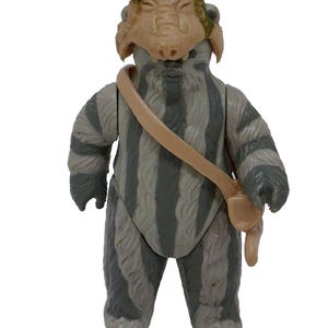 Może przedstawiać: Figurka akcji vintage postaci Ewoka z Gwiezdnych Wojen. Figurka ma szare i białe paski, beżową maskę i beżową torbę. Beżowa broń leży na dole. Figurka ma około 10 cm wysokości.