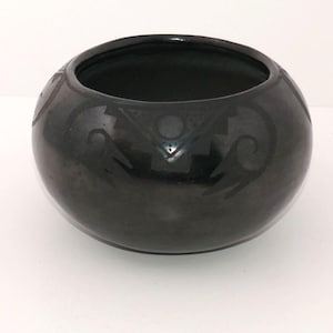 May include: A round, black ceramic bowl with a polished finish. The exterior features a matte black decorative band around the upper portion, displaying geometric patterns including triangles, circles, and curved lines in a subtle contrast.