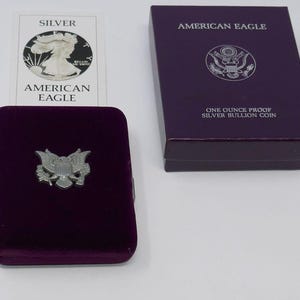 Może przedstawiać: Fioletowe aksamitne etui ze srebrnym emblematem orła, któremu towarzyszy pasujące fioletowe pudełko z napisem American Eagle One Ounce Proof Silver Bullion Coin oraz karta certyfikatu przedstawiająca monetę z napisem Silver American Eagle.