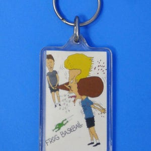 May include: A rectangular clear plastic keychain featuring a graphic illustration of two cartoon characters. One character has blonde hair and is hitting a brown object with a bat. The text FROG BASEBALL is printed in black below a green frog graphic.