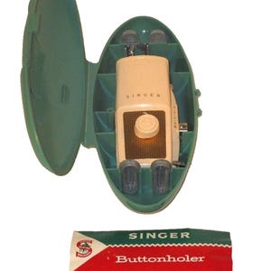 May include: Vintage Singer buttonholer machine in a green case. The beige buttonholer is inside the open case. The case is oval-shaped. A red, white, and green Singer Buttonholer box is below the case.