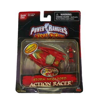 May include: A Power Rangers Ninja Storm Action Racer toy, featuring a red vehicle with gold and green accents, and a small red action figure. The packaging includes the text "Storm Megazord Action Racer".