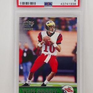 May include: A 2000 Pacific Tom Brady rookie football card, graded NM-MT 8 by PSA, encased in a protective holder. The card features Brady in his red and white uniform, holding a football. The card's text includes "2000 Pacific" and "Tom Brady".