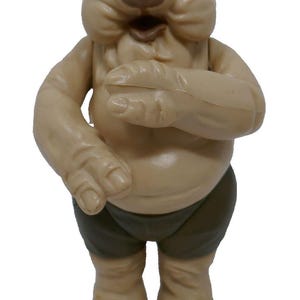 May include: A tan-colored action figure of a creature with a large head, small eyes, and a wide mouth. The figure has large hands and feet, and is wearing dark green shorts. The figure is standing upright.