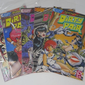 May include: A collection of five "Dirty Pair" comic books in plastic sleeves. The covers feature colorful anime-style illustrations of women, robots, and action scenes. The comic book titles are in bold, stylized fonts.