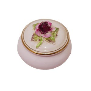 May include: A small, round, pink ceramic trinket box with a lid. The lid features a raised, detailed rose in shades of purple and pink, with green leaves. The rim of the lid and the base are trimmed with gold.