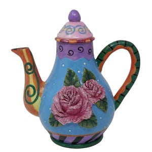 May include: A colorful ceramic teapot with a whimsical design. The teapot features a light blue body adorned with pink roses and green leaves. The handle and spout have orange and green accents, and the lid is topped with a purple knob.
