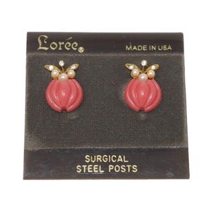 May include: A pair of pink earrings with a gold-colored accent and pearl and rhinestone details. The earrings are in the shape of a flower. The earrings are on a dark gray card that says "Loree" and "Made in USA". The card also says "Surgical Steel Posts".