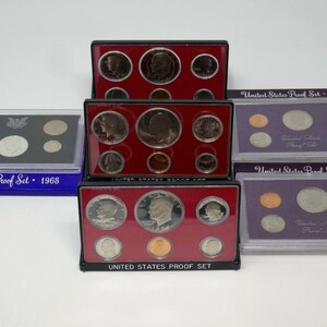 May include: Several United States Proof Sets, including sets from 1968, 1984, and 1985, are displayed. Each set contains multiple coins of varying sizes and colors, encased in protective plastic cases. The sets are in red and purple boxes.