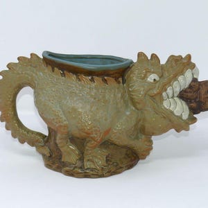 May include: A ceramic mug shaped like a green dragon with a textured body, a long tail forming the handle, and an open mouth revealing large white teeth. The interior of the mug is a smooth blue, and the dragon is biting down on a brown object.