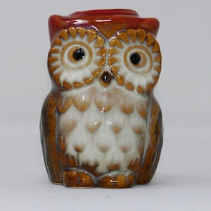 Może przedstawiać: Ceramiczna figurka sowy z błyszczącym szkliwem w odcieniach brązu, kremu i głębokiego czerwono-pomarańczowego koloru na górze. Sowa ma duże, teksturowane okrągłe oczy z ciemnymi źrenicami, mały dziób oraz wzór piór widoczny na jej klatce piersiowej.