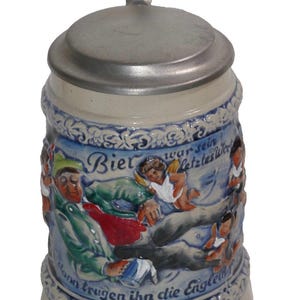 May include: A ceramic beer stein with a pewter lid and handle. The stein is decorated with a blue and white design featuring figures and text in German. The base is a light beige color.