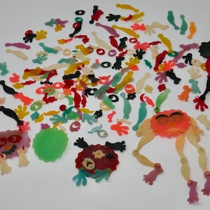 May include: A collection of colorful, whimsical plastic toy pieces scattered on a white surface. The pieces include various shapes, such as hands, circles, and abstract figures in shades of red, orange, yellow, green, and purple.