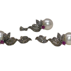May include: Three pieces of jewelry: a pair of earrings and a pendant. Each piece features a large, round, white pearl, a small red gemstone, and ornate silver-toned metalwork with leaf and marcasite details.