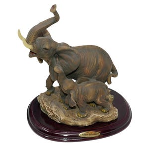 May include: A detailed figurine of two elephants, one adult and one calf, on a rocky base. The adult elephant has its trunk raised and tusks visible. The base is set on a dark brown, oval wooden stand. The figurine is labeled "Baby's Collection".